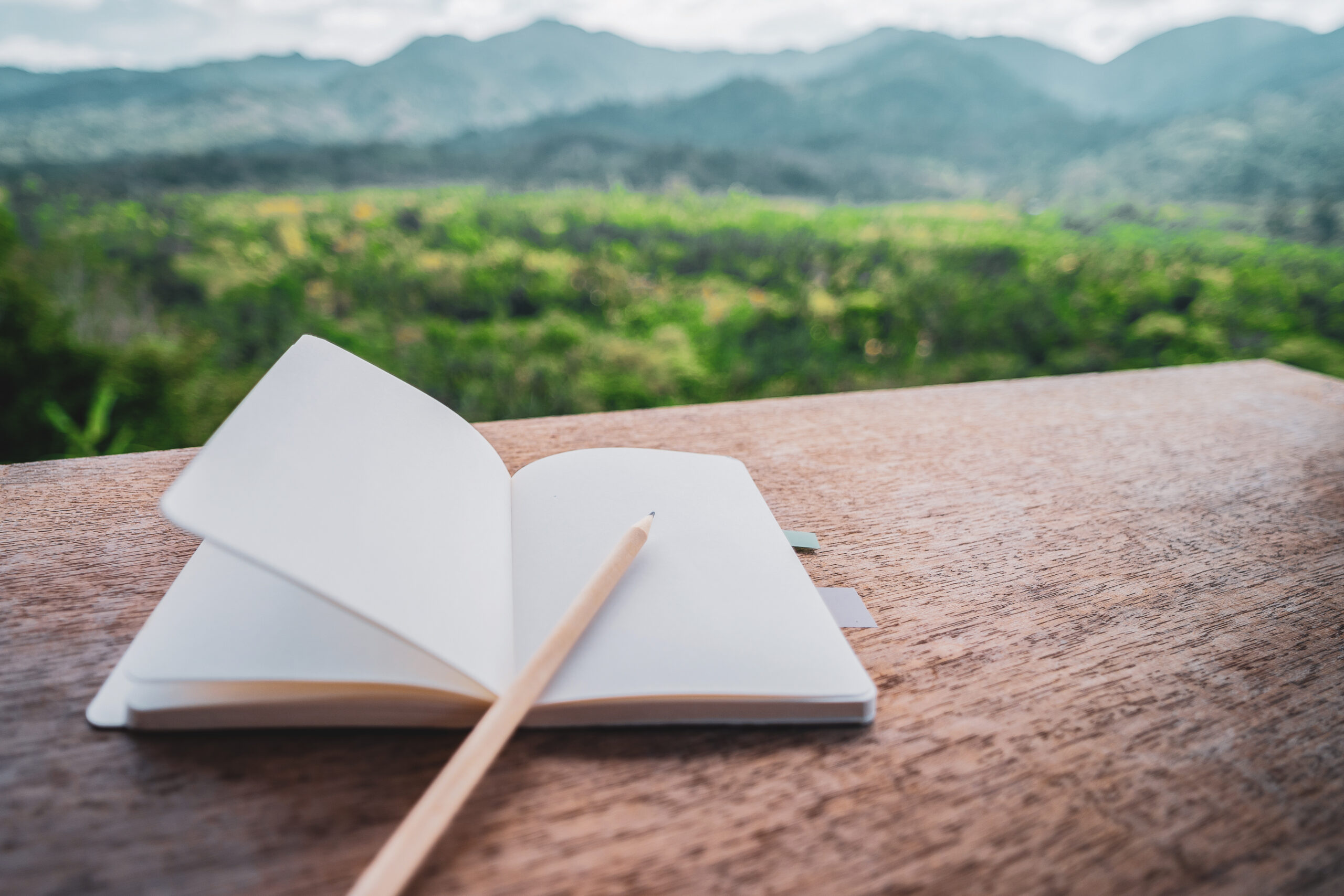 Notebook and pencil on wooden table with blur forrest and mountain background.